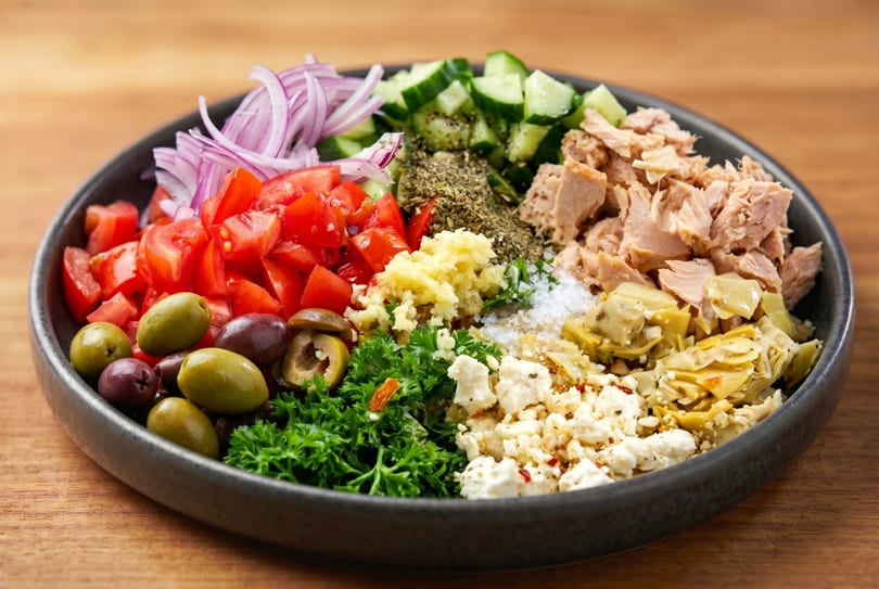 A vibrant salad bowl with tuna, tomatoes, olives, feta, artichokes, and herbs on a wooden table.