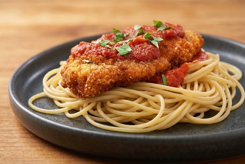 Crispy chicken parmesan with rich tomato sauce and fresh basil, served over spaghetti on a dark plate.