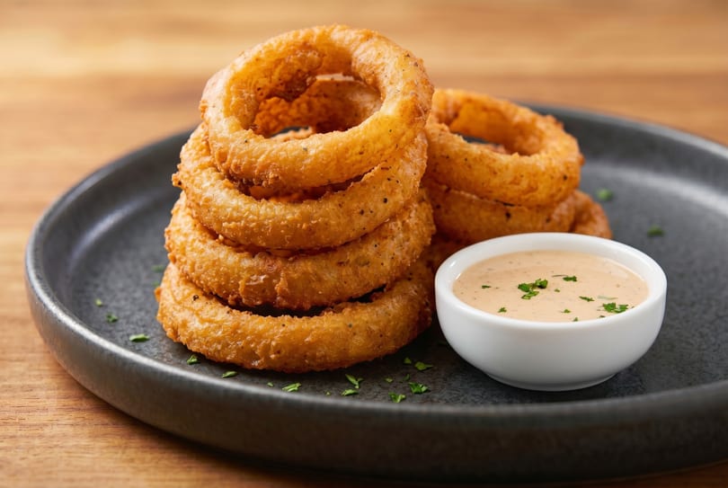 Golden-brown onion rings stacked on a dark plate with a side of dipping sauce.