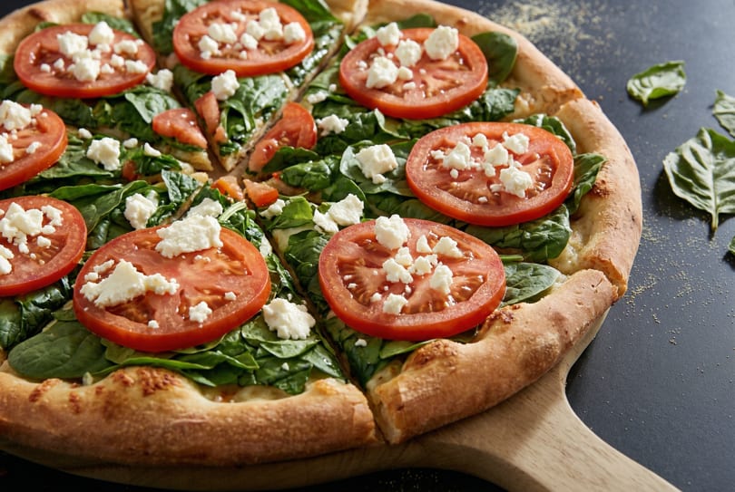 A freshly baked pizza with spinach, sliced tomatoes, and crumbled feta cheese on a wooden peel.