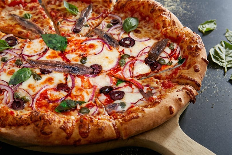 A freshly baked pizza with anchovies, red onion, olives, capers, and basil on a wooden peel.