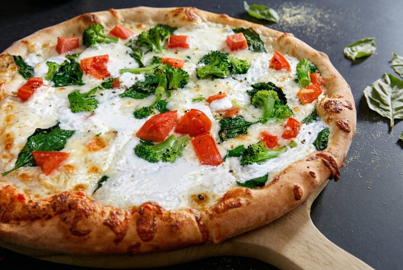 White pizza with broccoli, tomatoes, and spinach on a wooden peel.
