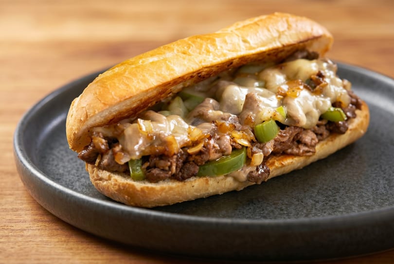 Cheesesteak sandwich with melted cheese, steak, grilled onions, and green peppers on a dark plate.