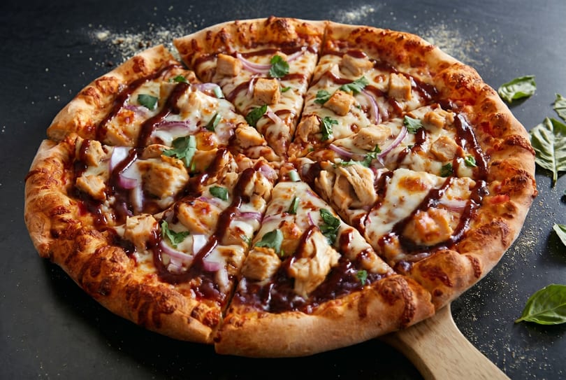 A whole BBQ chicken pizza, sliced, with red onion, cilantro, and a drizzle of sauce.