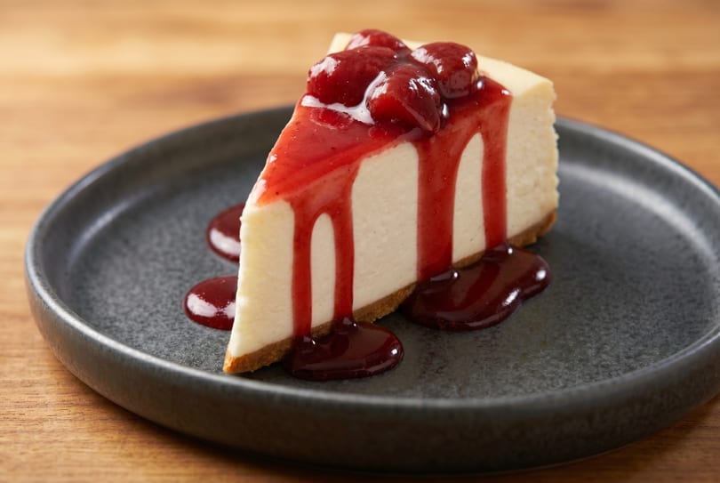 Slice of cheesecake with strawberry topping and sauce on a dark plate.