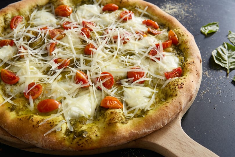 Pesto pizza with cherry tomatoes, fresh mozzarella, and shredded Parmesan on a wooden peel.